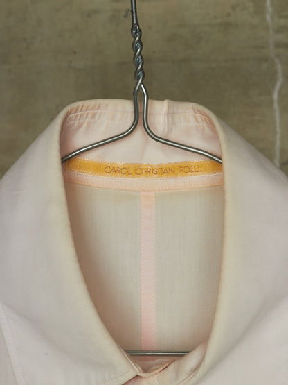 A close-up of a pale blush pink fabric garment hanging on a metal hanger against a textured concrete wall. The inner collar band is visible with the brand name 'CAROL CHRISTIAN TOELL' printed in gold.