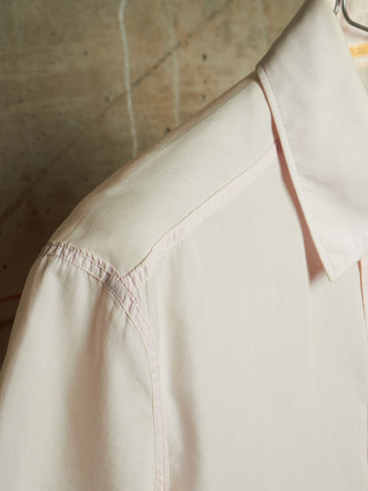 A close-up of a light cream or off-white button-up shirt on a hanger, showing the collar, shoulder seam, and sleeve against a textured concrete wall background.