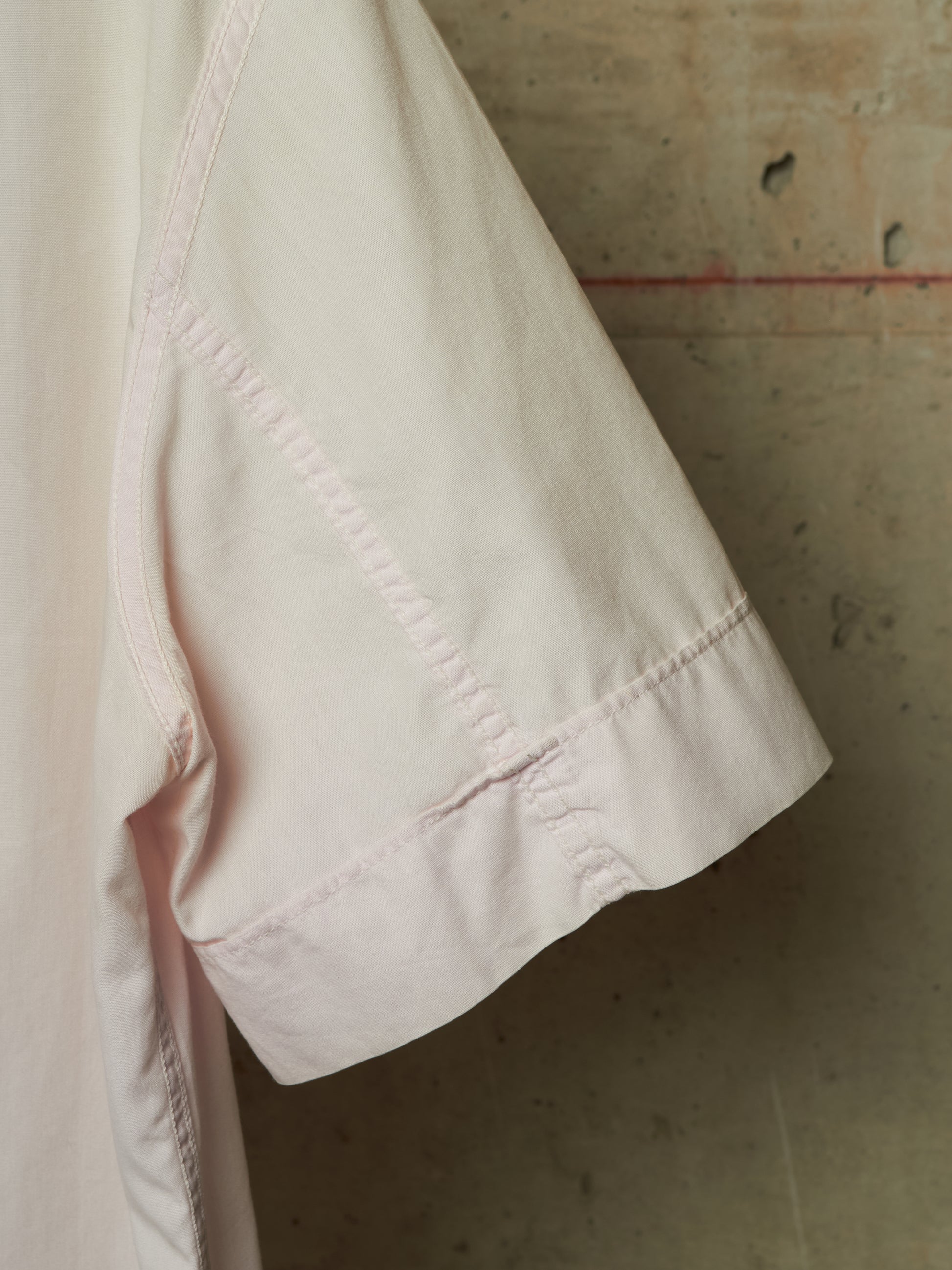 Close-up detail of a light pink short-sleeve garment, showing the sleeve cuff with decorative contrast stitching against a textured concrete wall background.
