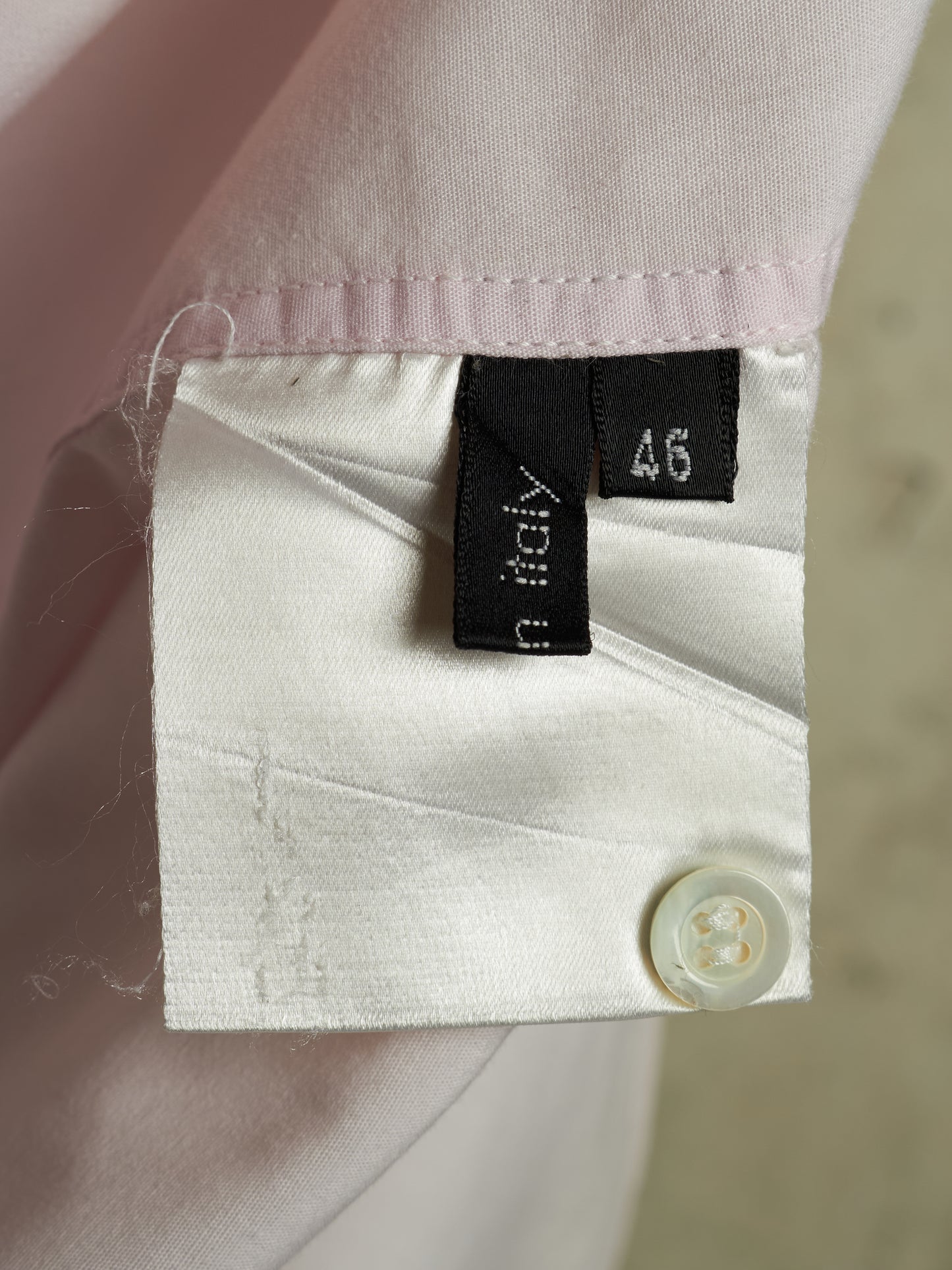 Close-up of a light pink dress shirt cuff with a white button, showing the inner lining and a black fabric brand tag that reads 'in italy' alongside a size tag reading '46'.