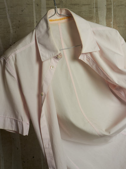 A pale blush pink short-sleeve button-up shirt with matching pink contrast stitching, hanging on a wire hanger against a textured concrete wall.