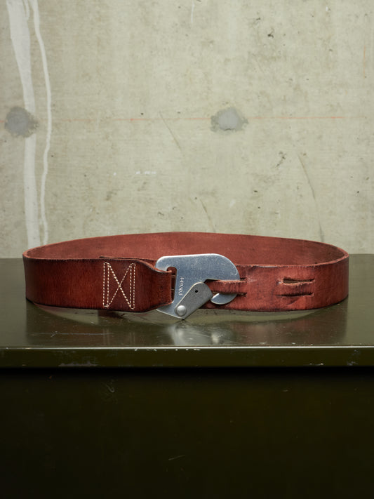 A reddish-brown leather belt with a silver-toned metal buckle, resting on a dark green surface against a textured concrete wall.