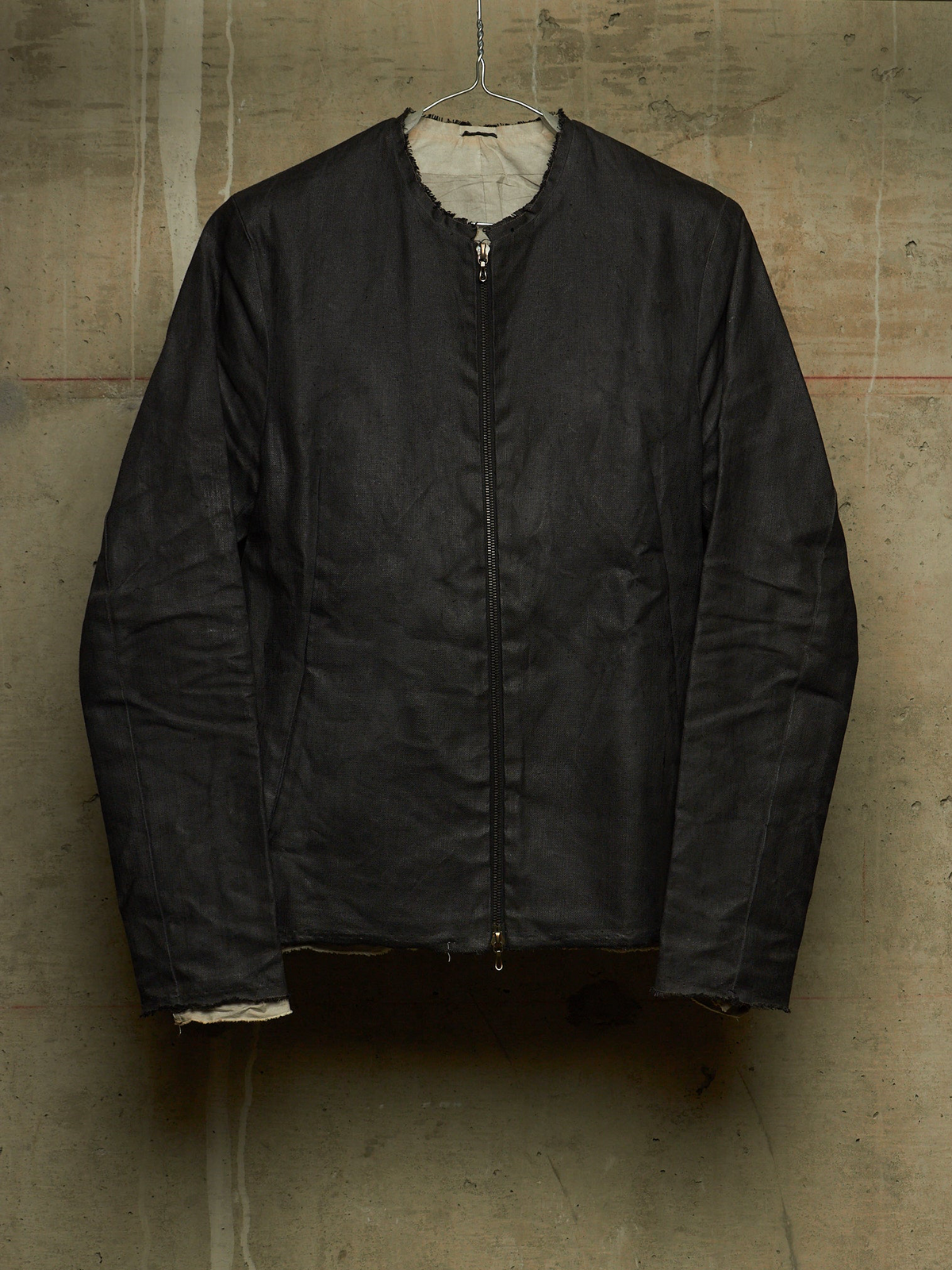 A black zip-up jacket with a light beige lining, hanging on a wire hanger against a textured concrete wall.