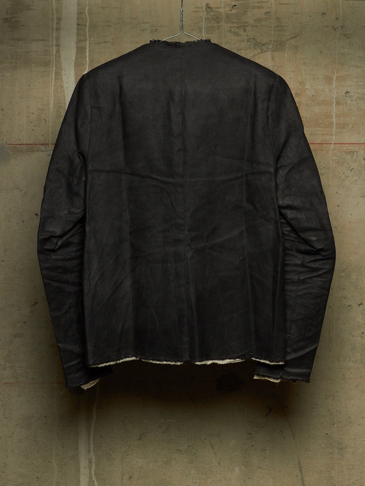 A black long-sleeve jacket or overshirt hanging against a textured concrete wall, showing the back view with raw, frayed hems.
