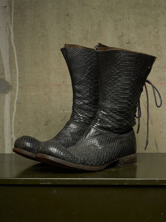 A pair of black snakeskin-textured mid-calf boots with side laces, displayed on a dark green surface against a textured concrete wall.