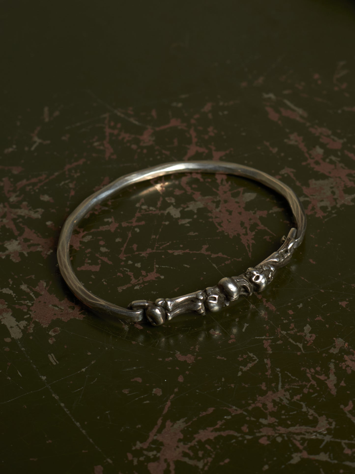 A polished silver-toned metal bangle bracelet featuring a decorative cluster of small skull motifs along the front band, displayed on a dark, textured background.