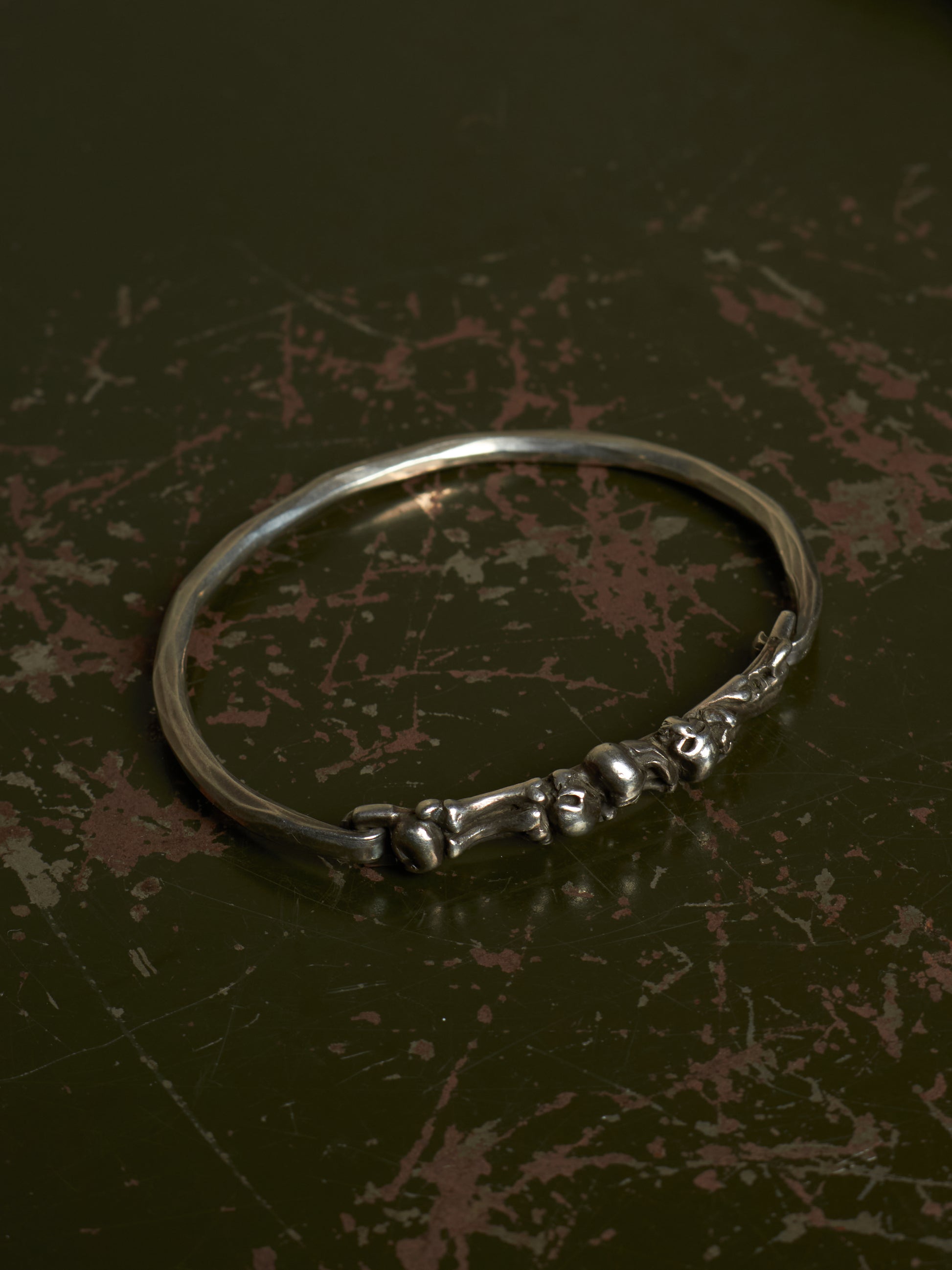 A polished silver-toned metal bangle bracelet featuring a decorative cluster of small skull motifs along the front band, displayed on a dark, textured background.