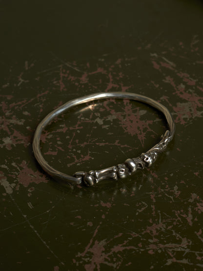 A polished silver-toned metal bangle bracelet featuring a decorative cluster of small skull motifs along the front band, displayed on a dark, textured background.