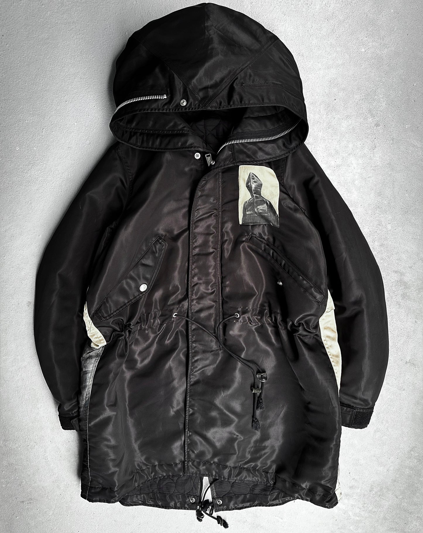 The UNDERCOVER AW16 ‘Borremans’ Full-zip Fishtail Nylon Parka by Undercover is a black hooded jacket with light side panels, front zipper, snap buttons, adjustable drawstrings, and a hooded figure patch on the chest.