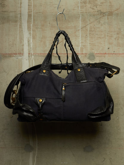 A dark navy blue canvas duffel bag with black leather handles and trim, hanging from a metal hanger against a textured concrete wall.