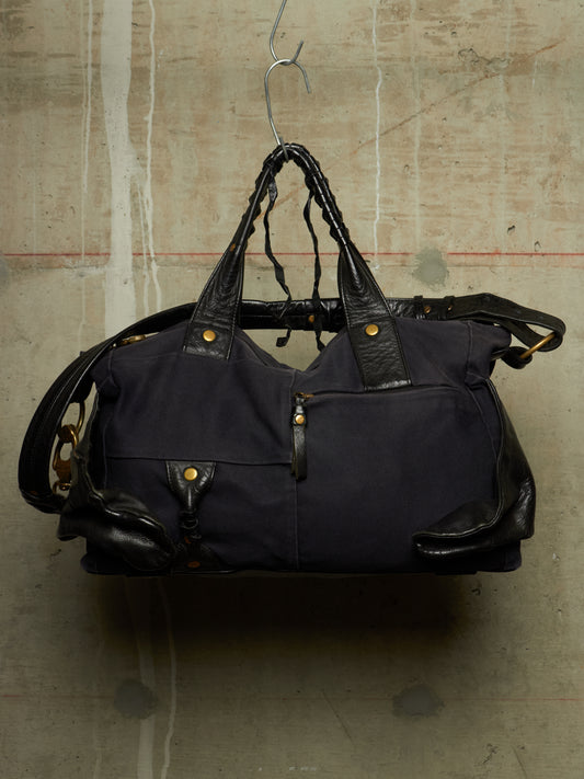 A dark navy blue canvas duffel bag with black leather handles and trim, hanging from a metal hanger against a textured concrete wall.