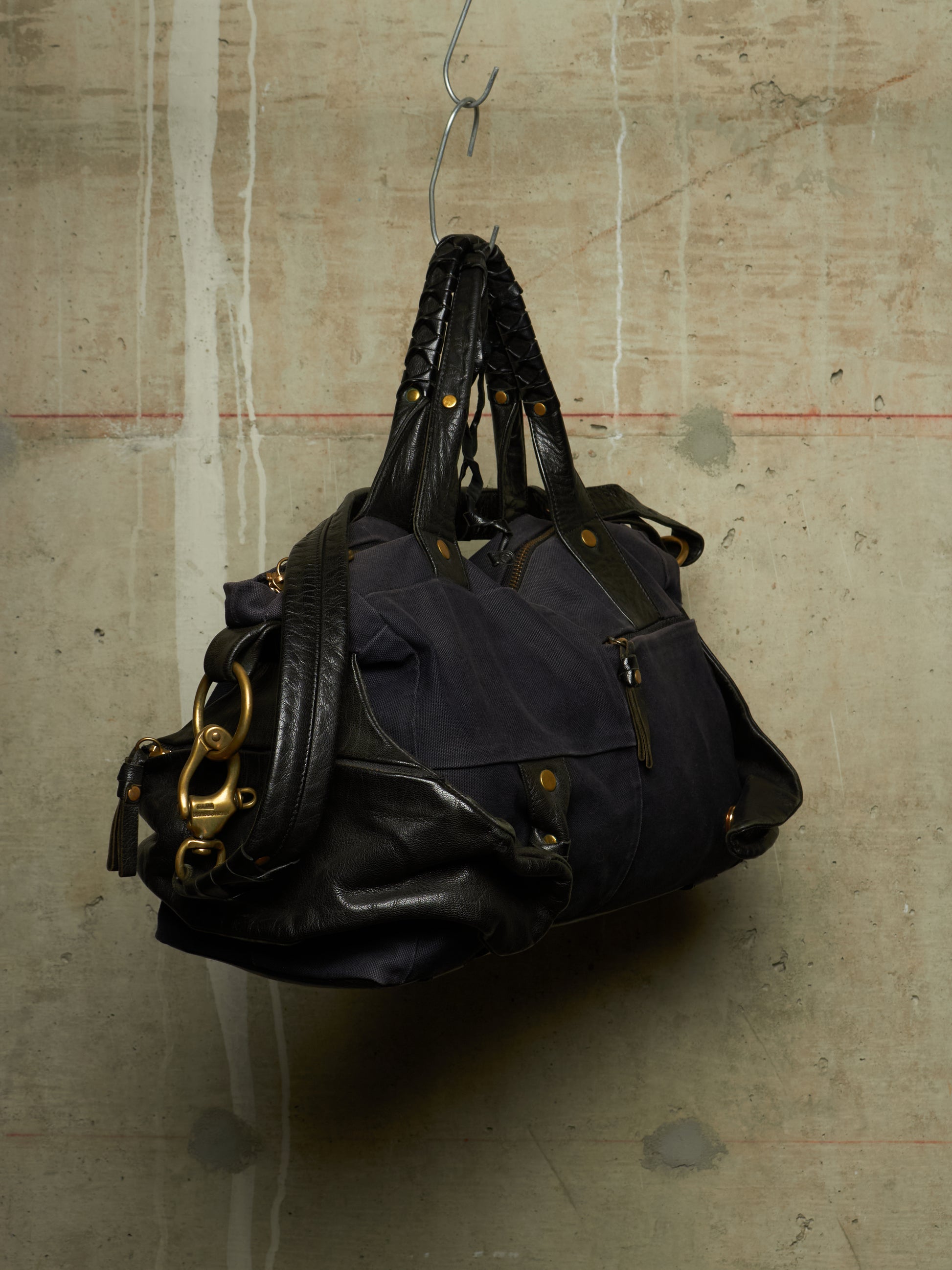 A dark navy blue and black leather handbag with gold-tone hardware, hanging from a metal hook against a textured concrete wall.