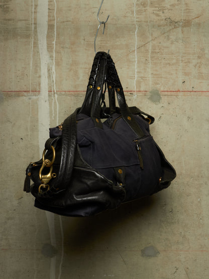 A dark navy blue and black leather handbag with gold-tone hardware, hanging from a metal hook against a textured concrete wall.