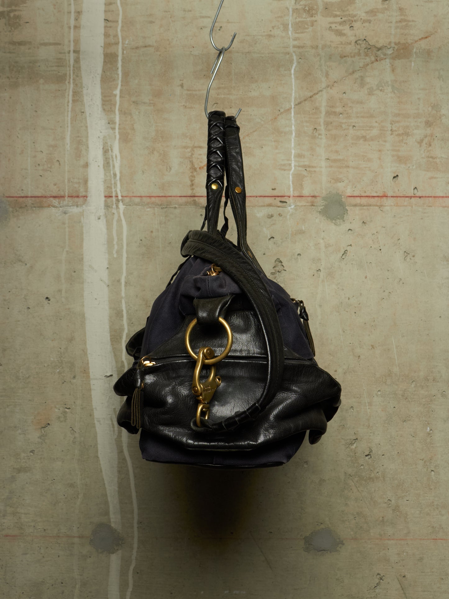 A black leather shoulder bag with gold hardware and a braided top handle, hanging from a metal hook against a textured concrete wall.