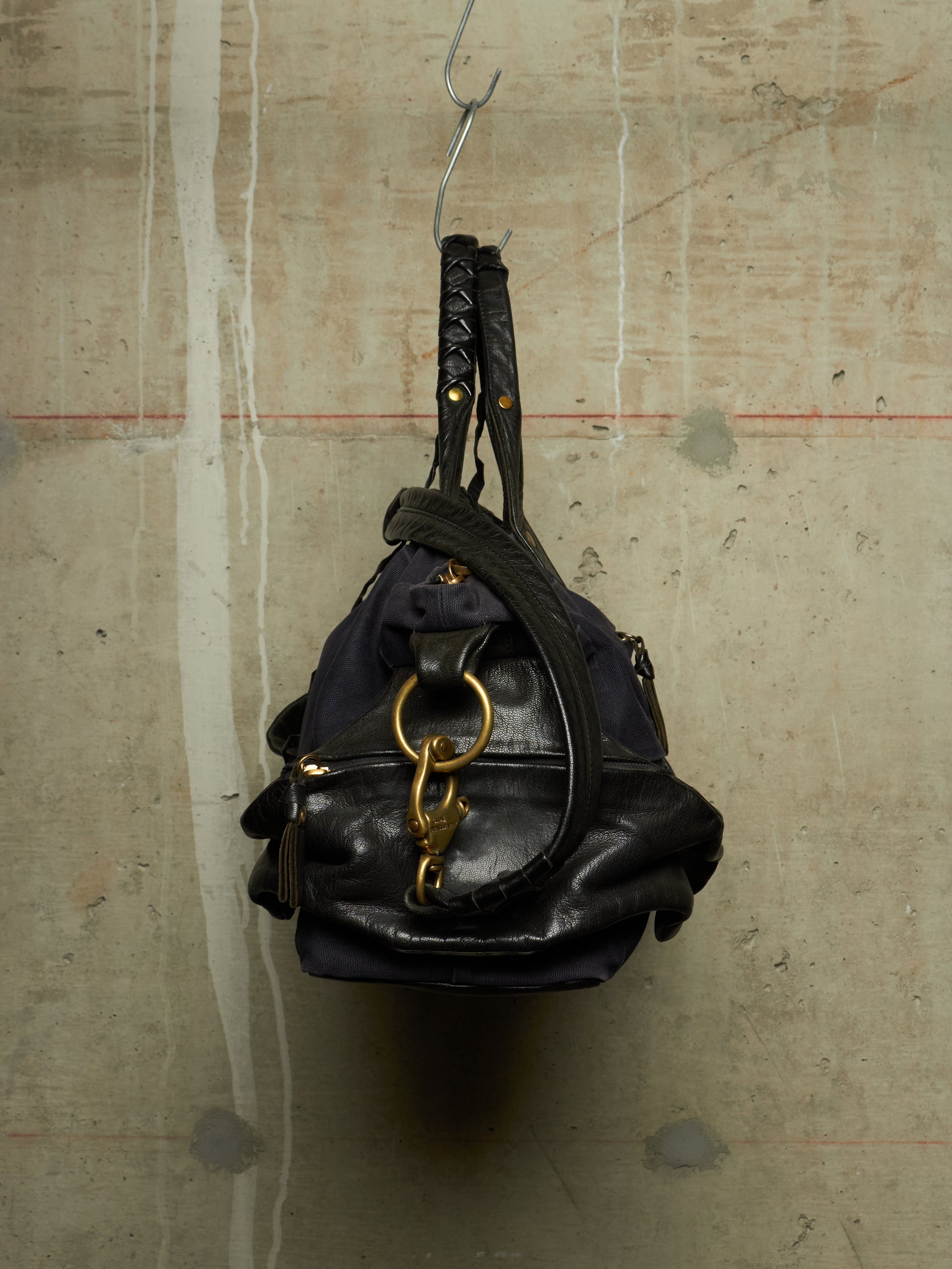 A black leather shoulder bag with gold hardware and a braided top handle, hanging from a metal hook against a textured concrete wall.