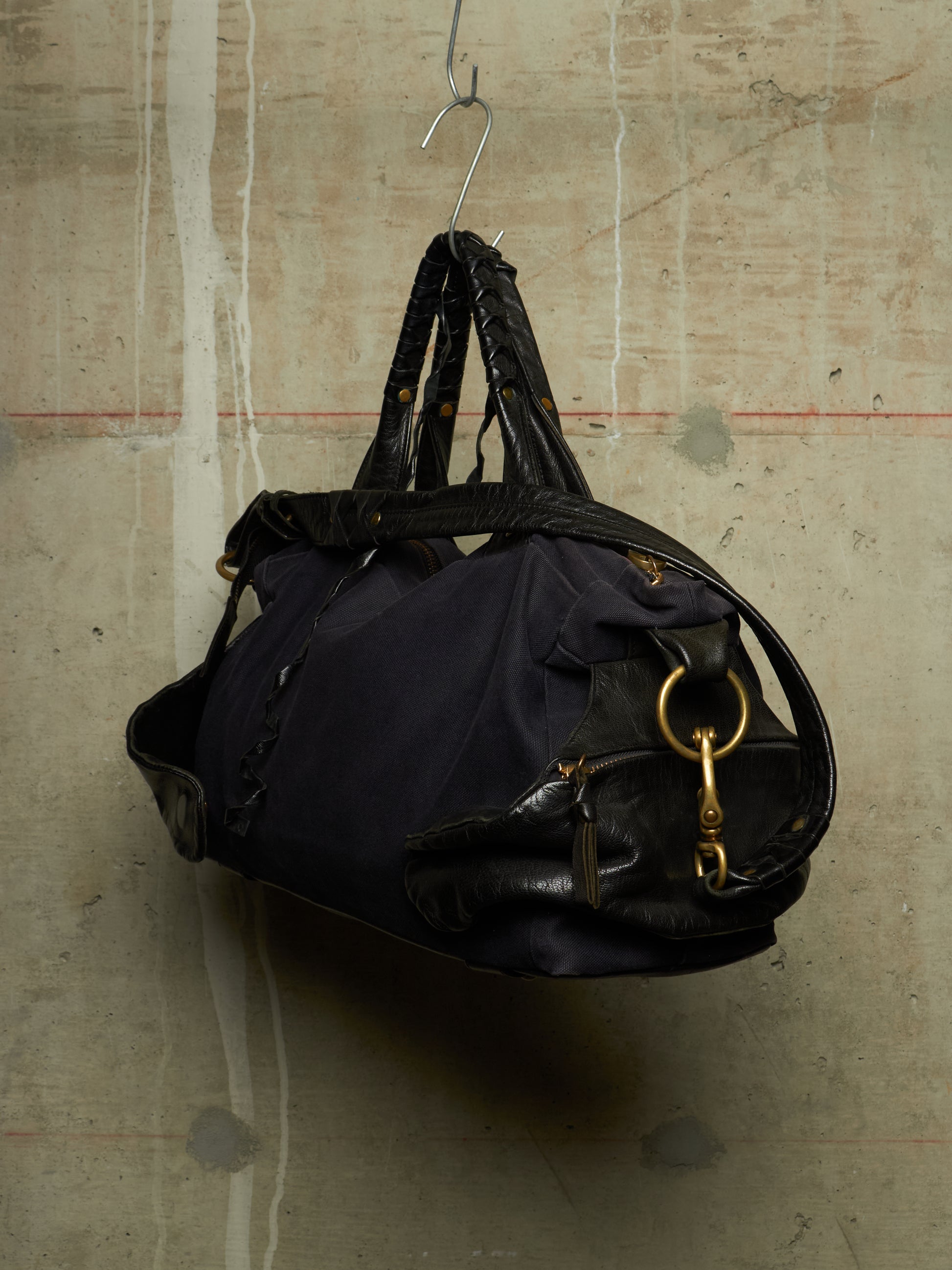 A dark navy blue nylon duffel bag with black leather trim and handles, hanging from a metal hook against a textured concrete wall.