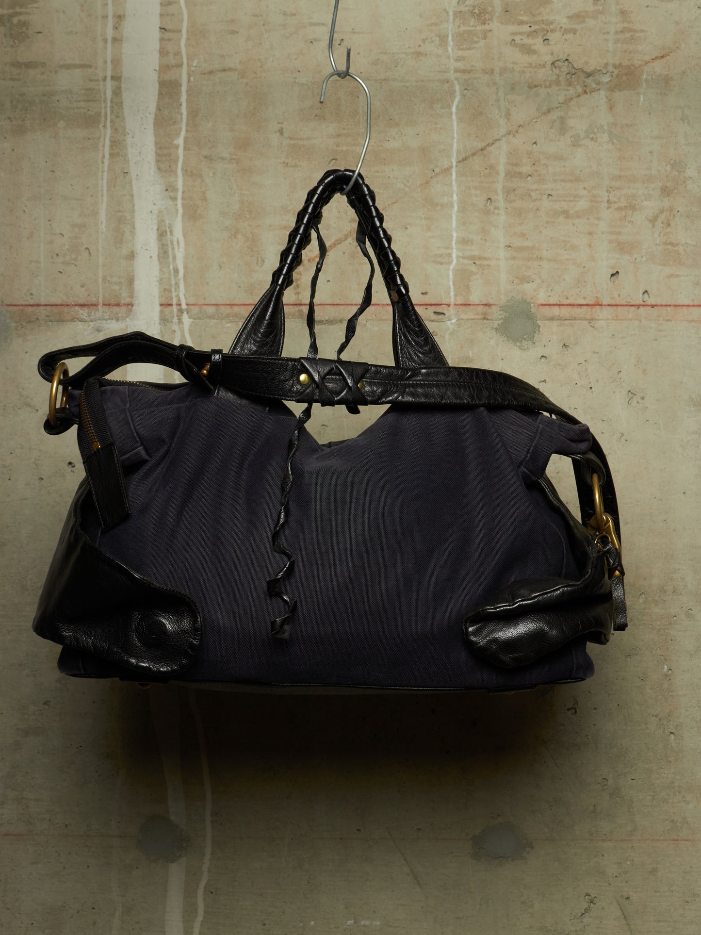 A dark navy blue nylon duffel bag with black leather trim, braided top handles, and gold-tone hardware, hanging from a metal hanger against a textured concrete wall.