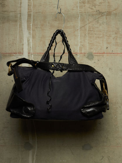 A dark navy blue nylon duffel bag with black leather trim, braided top handles, and gold-tone hardware, hanging from a metal hanger against a textured concrete wall.