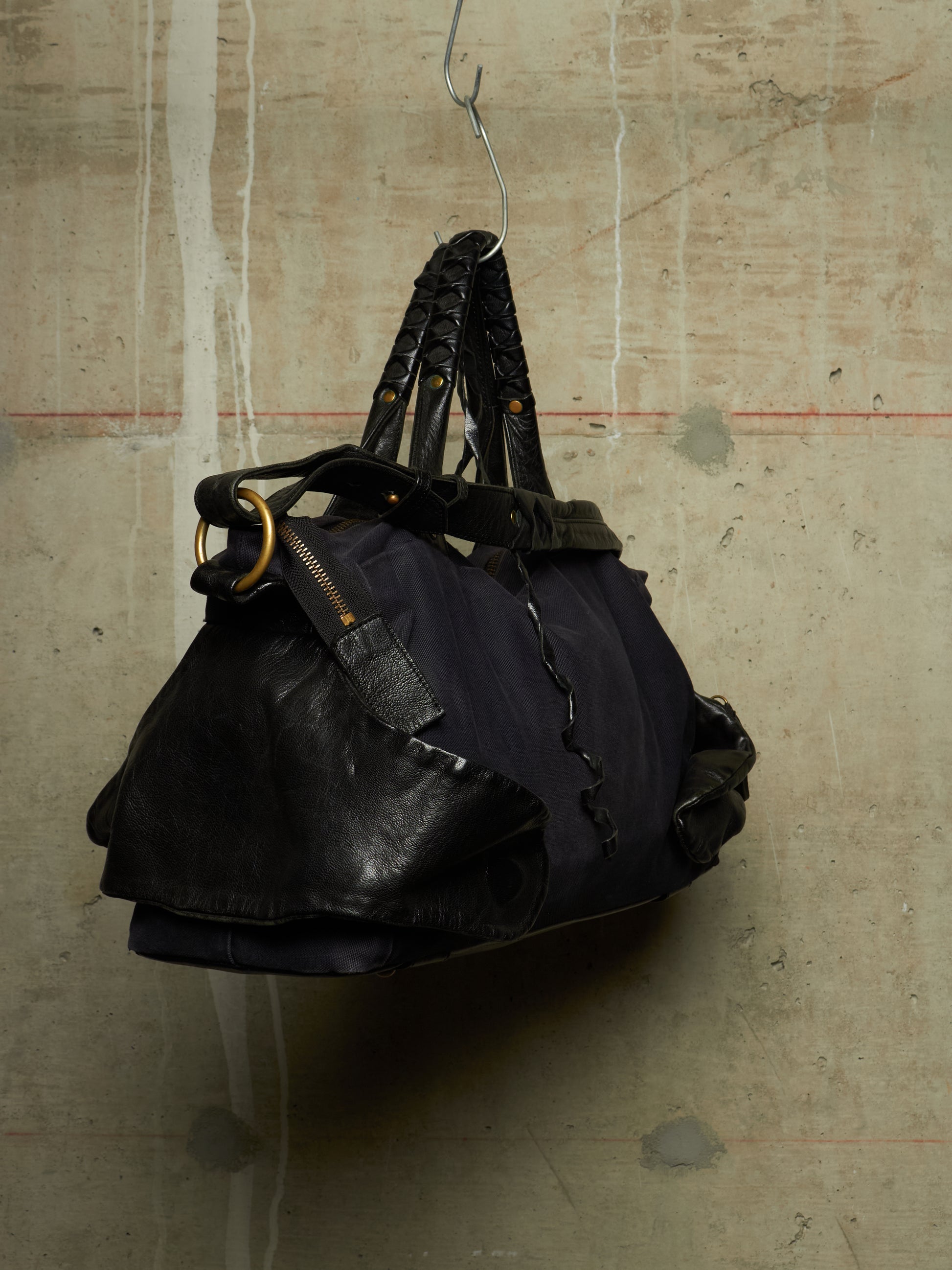 A black leather and fabric duffel bag with braided shoulder straps and gold-tone hardware, hanging from a metal hook against a textured concrete wall.