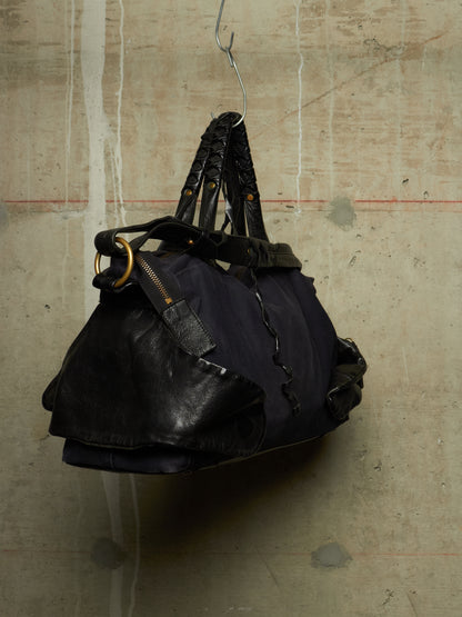 A black leather and fabric duffel bag with braided shoulder straps and gold-tone hardware, hanging from a metal hook against a textured concrete wall.