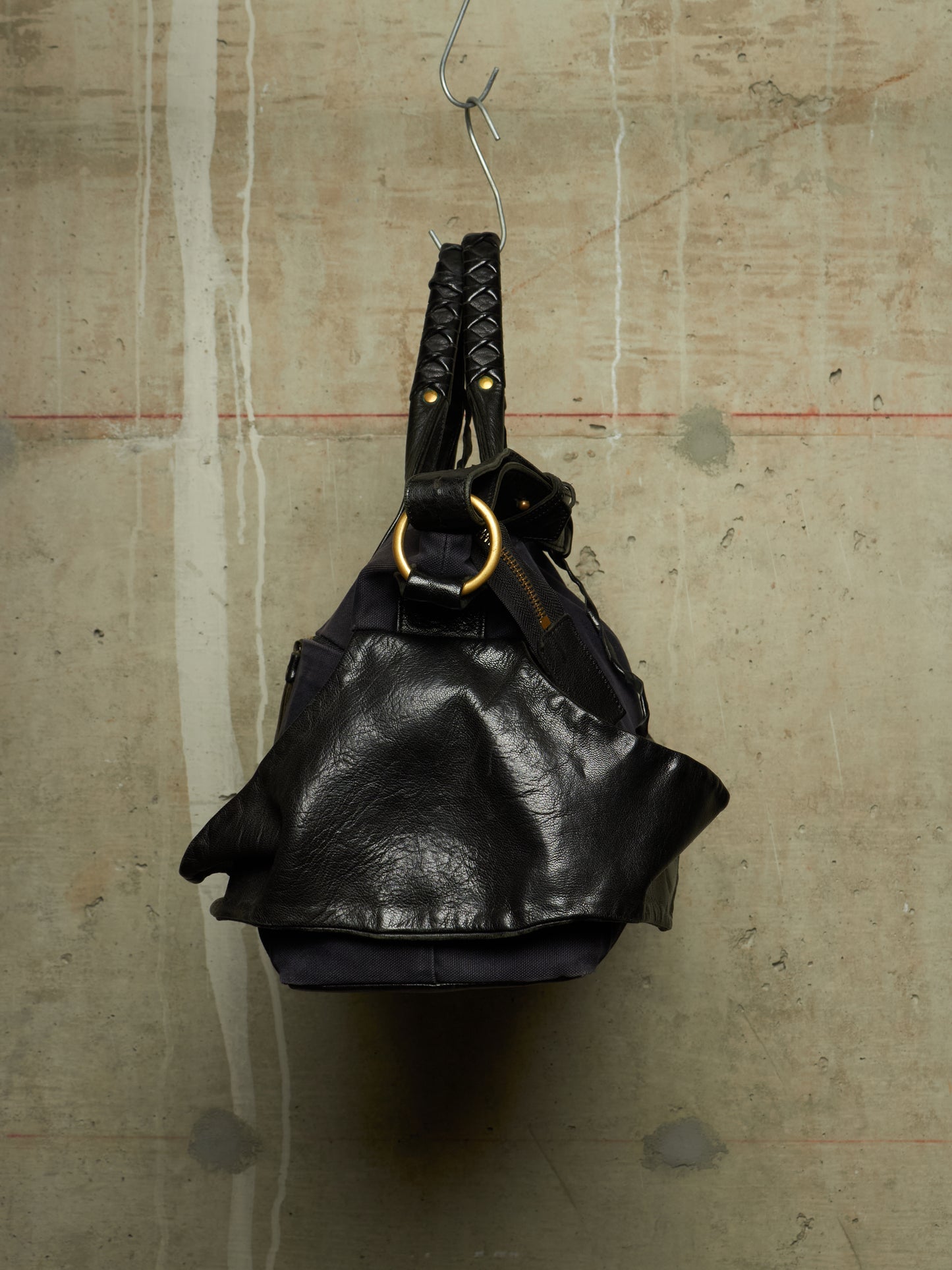 A black leather shoulder bag with braided handles and a large gold-tone ring detail, hanging from a metal hook against a textured concrete wall.