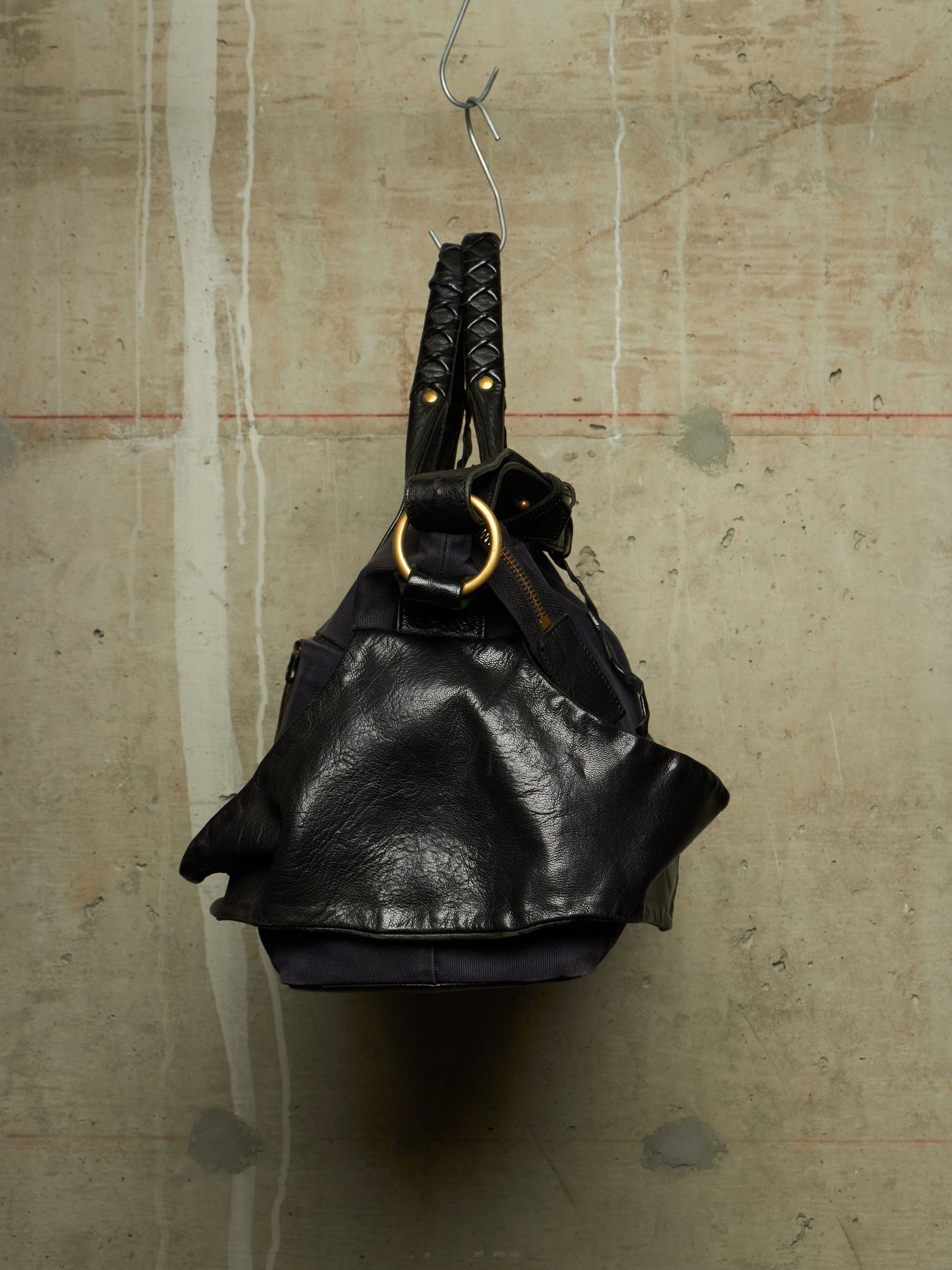 A black leather shoulder bag with braided handles and a large gold-tone ring detail, hanging from a metal hook against a textured concrete wall.