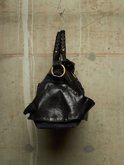 A black leather shoulder bag with braided handles and a large gold-tone ring detail, hanging from a metal hook against a textured concrete wall.