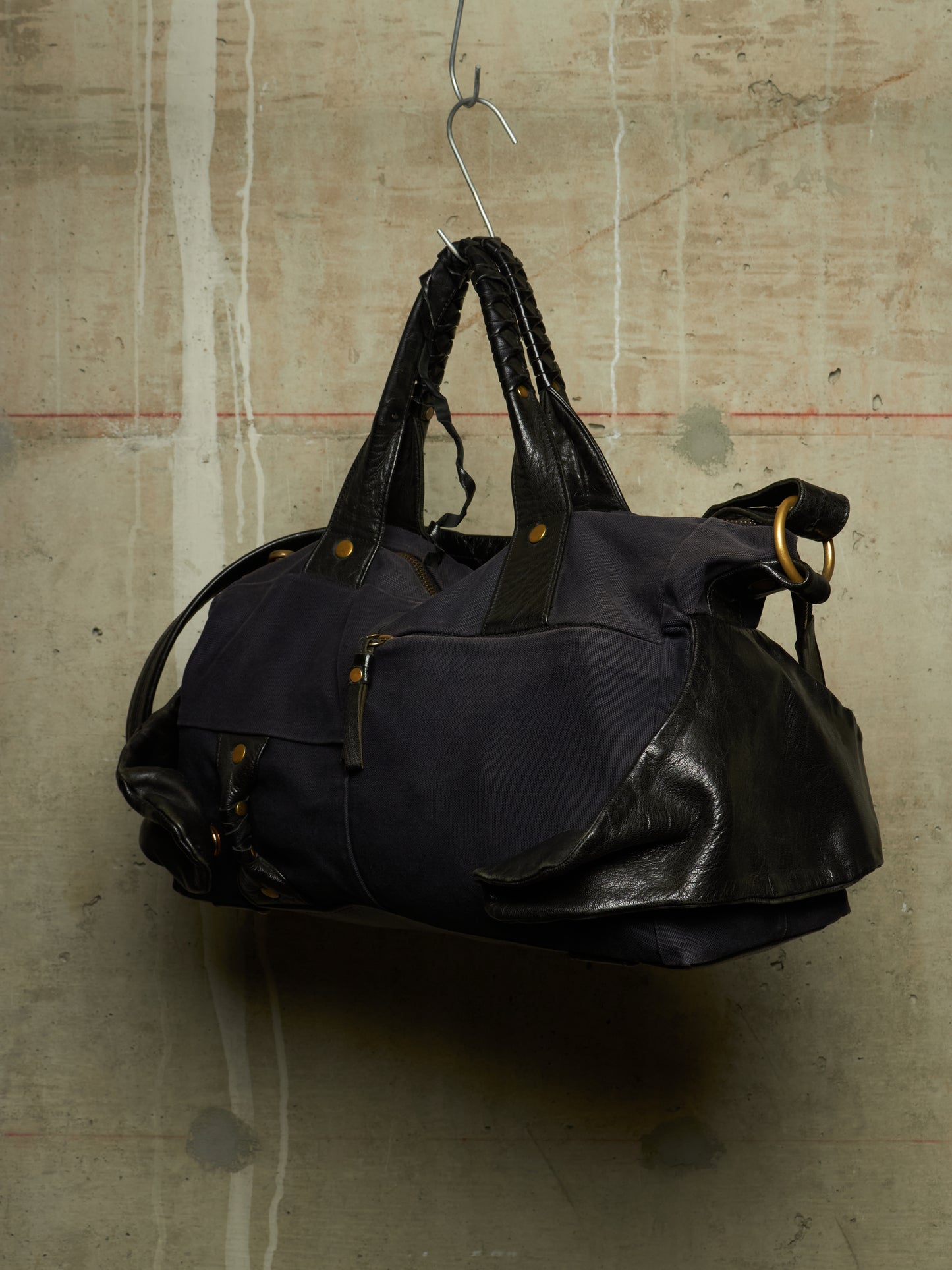 A dark navy blue duffel bag with black leather handles and trim, hanging from a metal hook against a textured concrete wall.