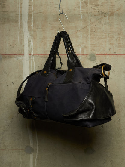 A dark navy blue duffel bag with black leather handles and trim, hanging from a metal hook against a textured concrete wall.