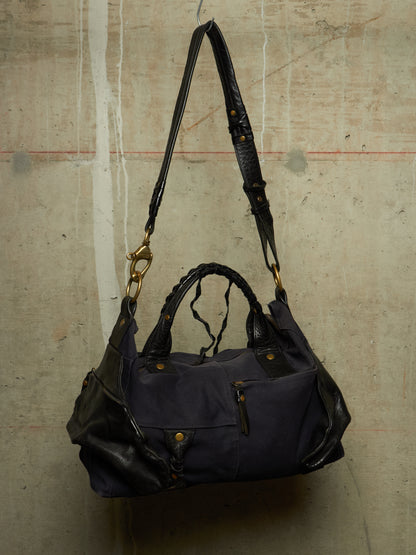 A navy blue nylon shoulder bag with black leather trim, top handles, and a long adjustable shoulder strap, hanging against a textured concrete wall.