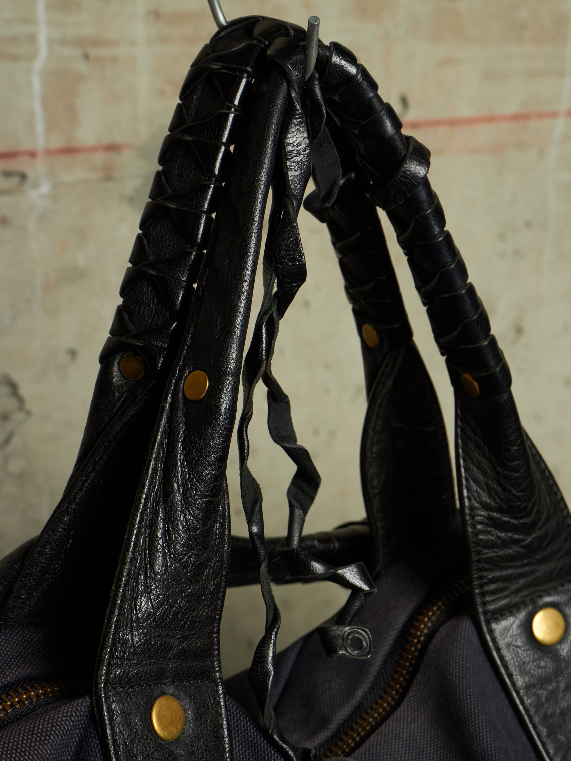 Close-up of a black leather shoulder bag with braided leather handles, brass-tone rivets, and a dark fabric body, photographed against a textured beige wall.
