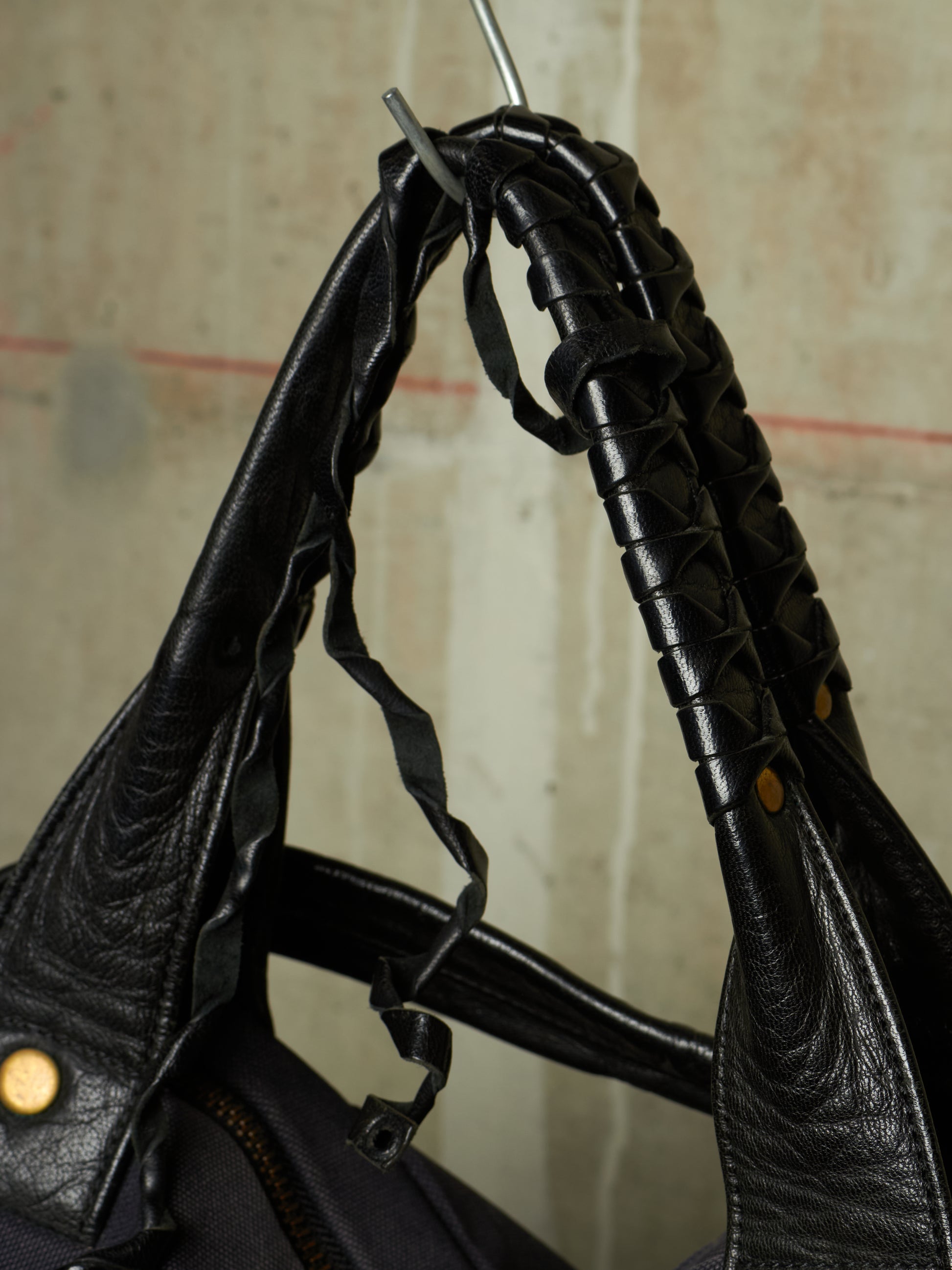 Close-up of a black leather shoulder bag with textured, woven, and stitched details on the handle, hanging against a neutral concrete wall.