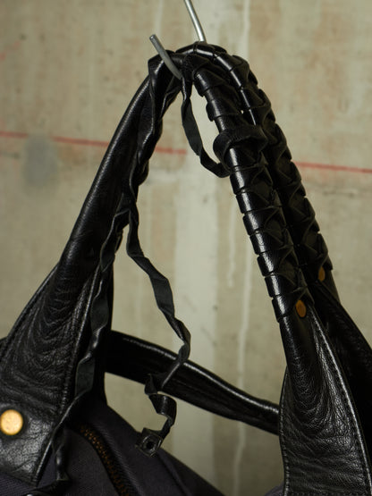Close-up of a black leather shoulder bag with textured, woven, and stitched details on the handle, hanging against a neutral concrete wall.