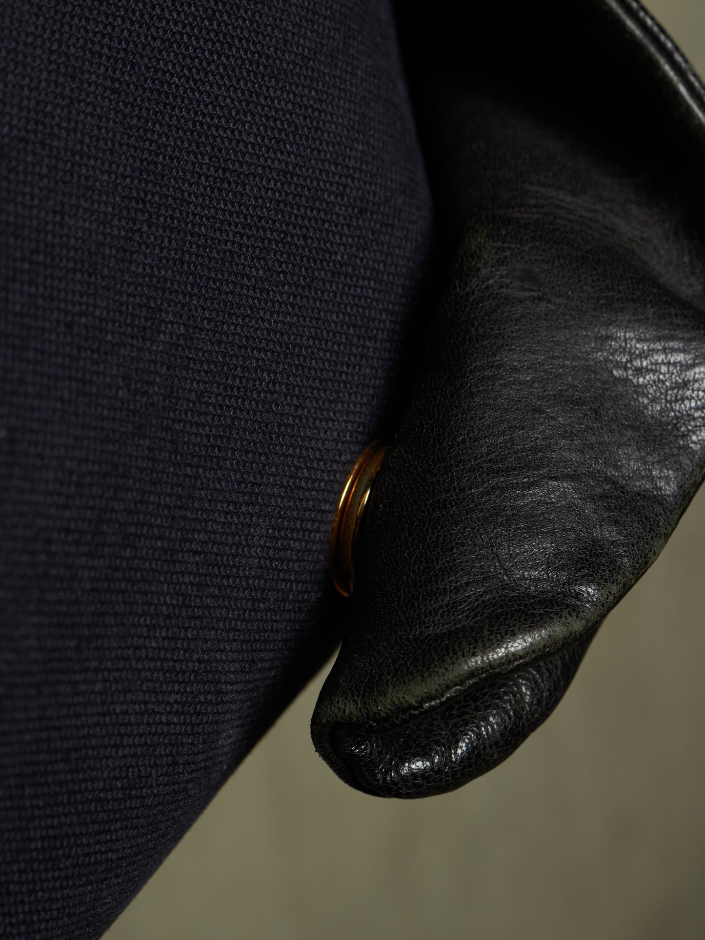 Close-up detail of a black leather shoulder strap resting against a dark navy blue woven fabric, with a small gold-toned metal ring visible.