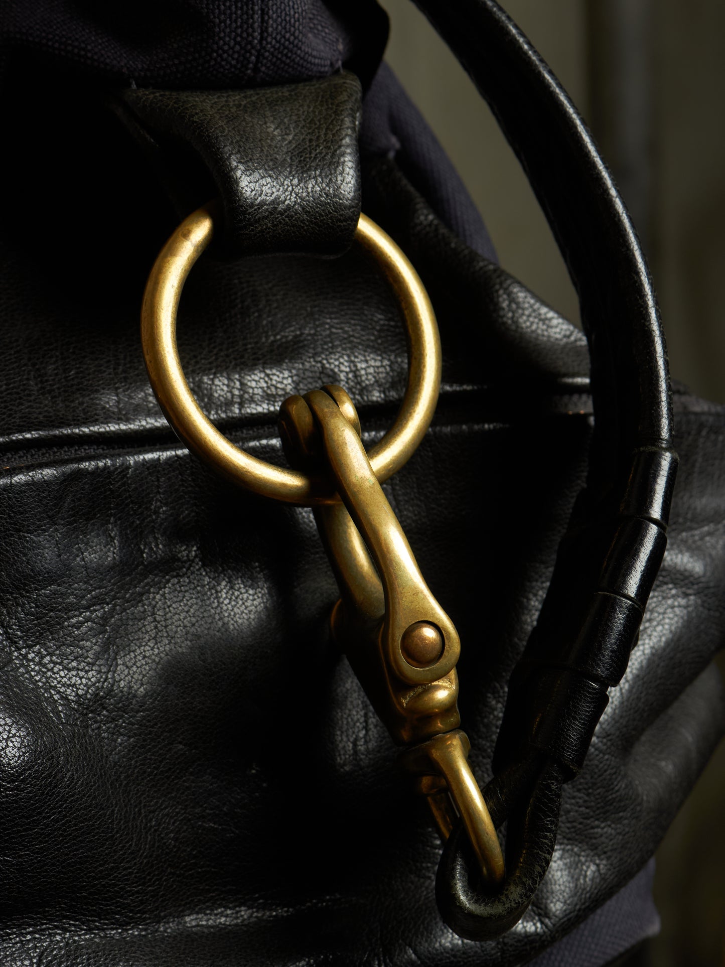 Close-up of a black leather bag featuring a large brass ring and matching brass snap hook hardware, with a dark fabric strap visible in the background.