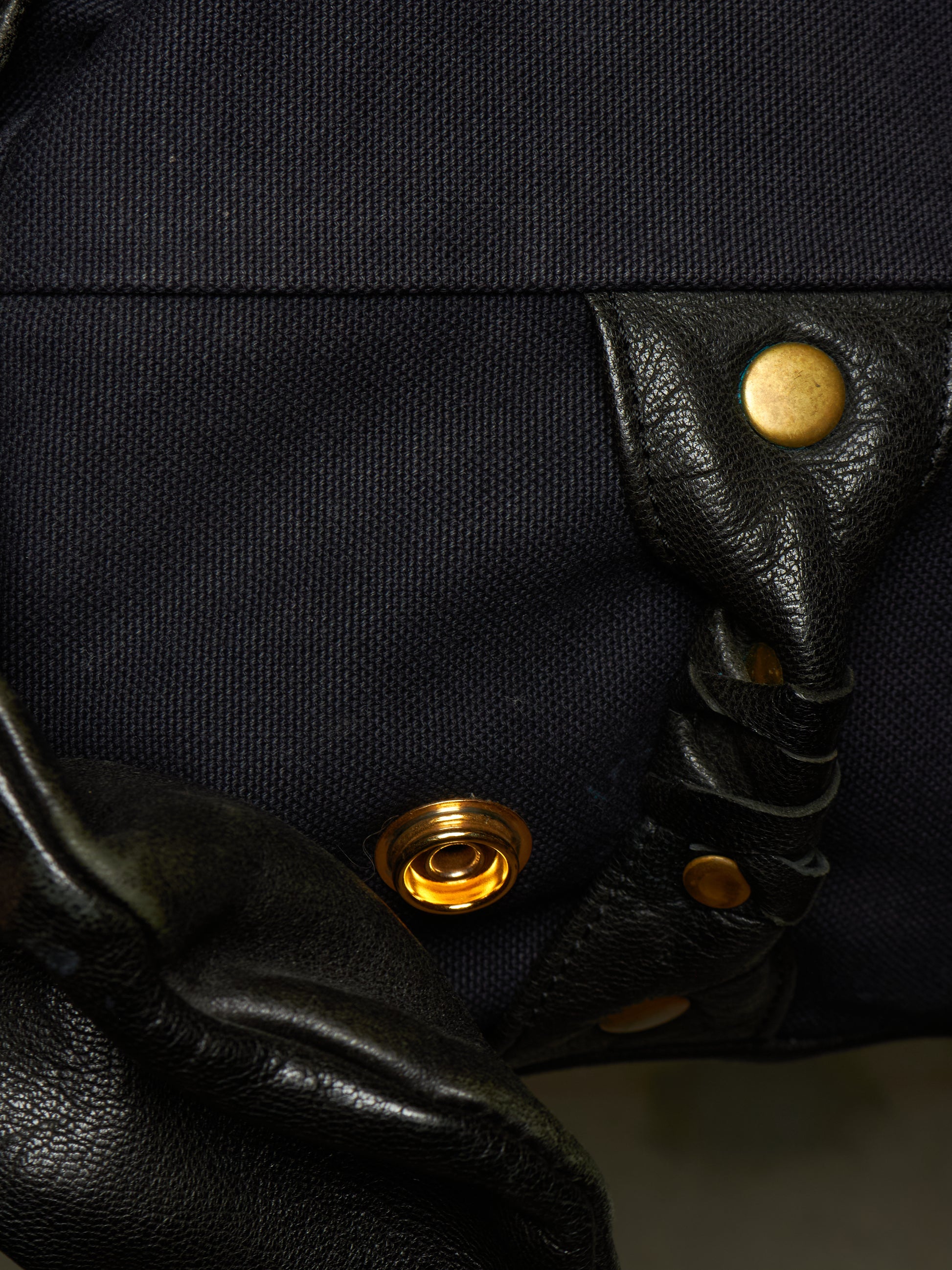Close-up detail of a black canvas and leather bag featuring brass snap buttons and a braided leather strap.