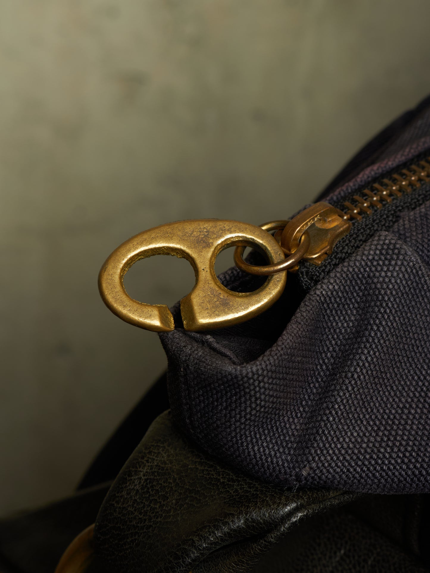 Close-up of a dark navy or black textured fabric bag with a prominent brass zipper pull and matching ring hardware, set against a neutral gray background.