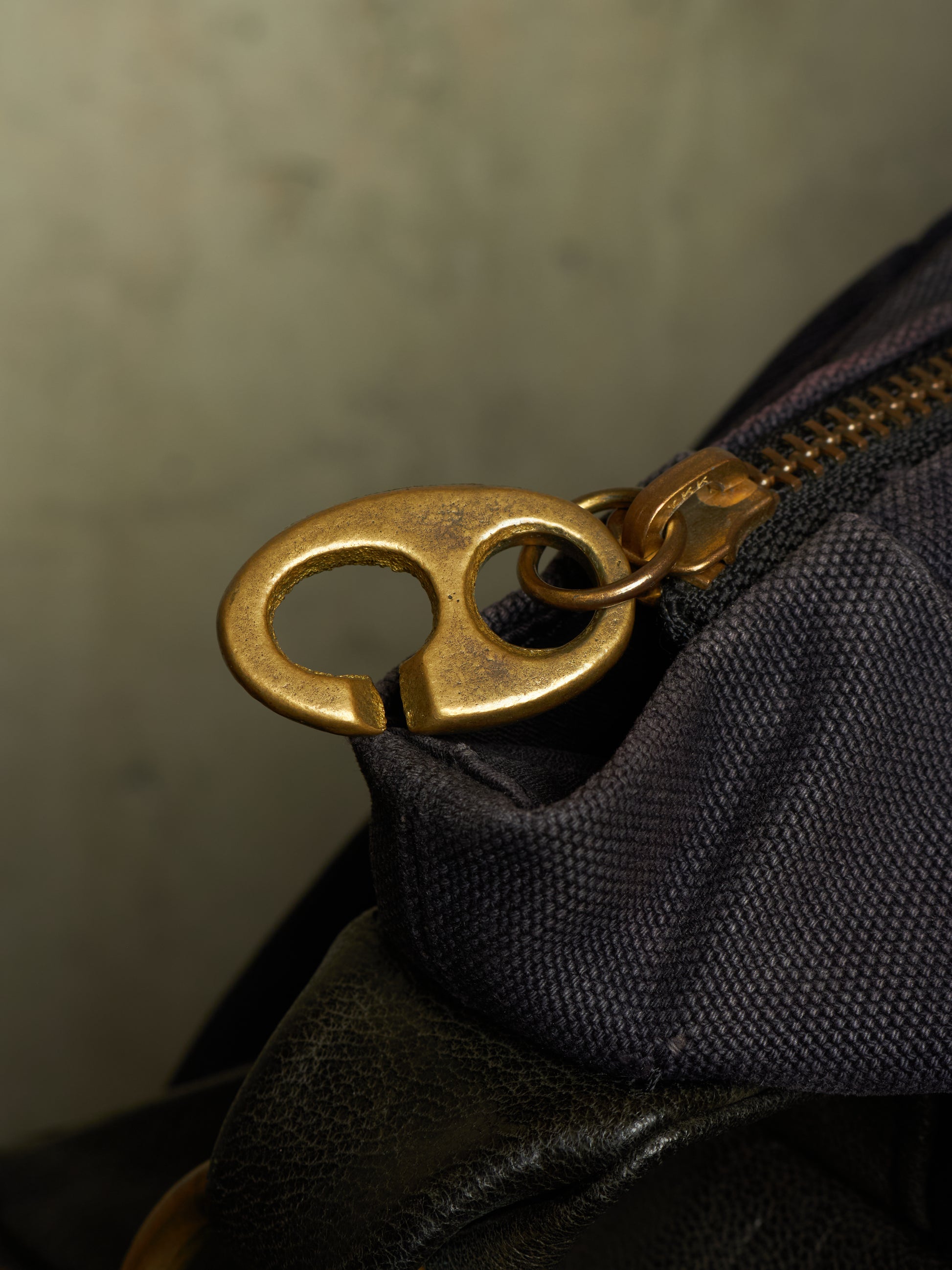 Close-up of a dark navy or black textured fabric bag with a prominent brass zipper pull and matching ring hardware, set against a neutral gray background.