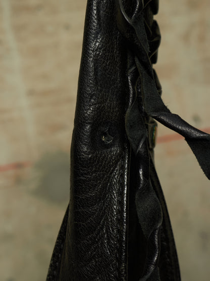 Close-up of a black leather bag or garment showing textured leather, a small button, and a dark ribbon detail against a blurred beige background.