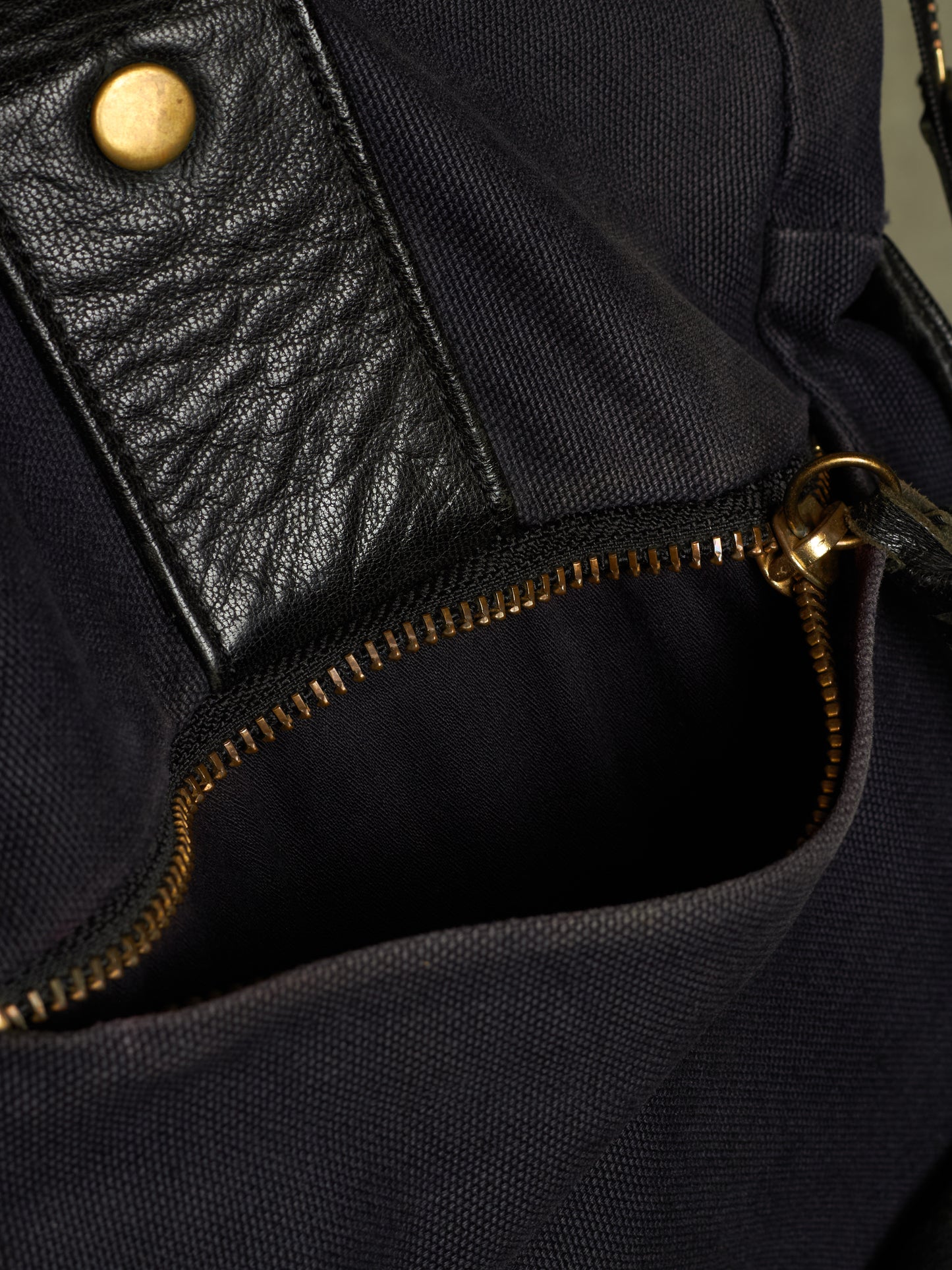 Close-up of a dark navy canvas bag with a black leather pocket and gold-tone metal hardware, including a zipper and snap button.