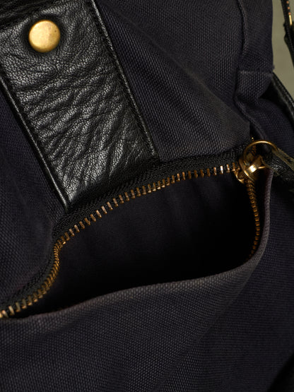 Close-up of a dark navy canvas bag with a black leather pocket and gold-tone metal hardware, including a zipper and snap button.