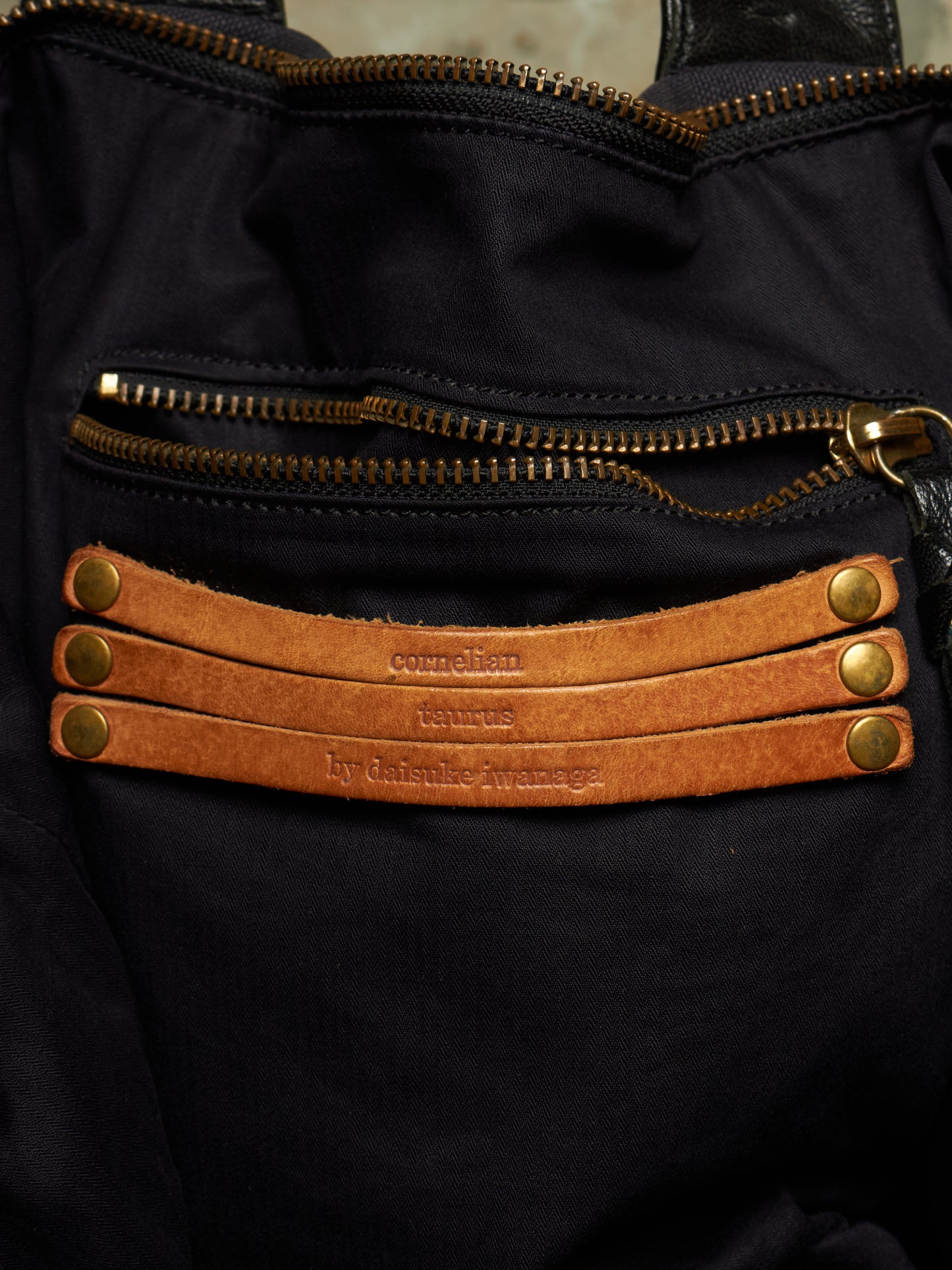 Close-up of a black fabric bag with gold-tone zippers and three tan leather straps across the front, each stamped with the brand name 'corbelian'.