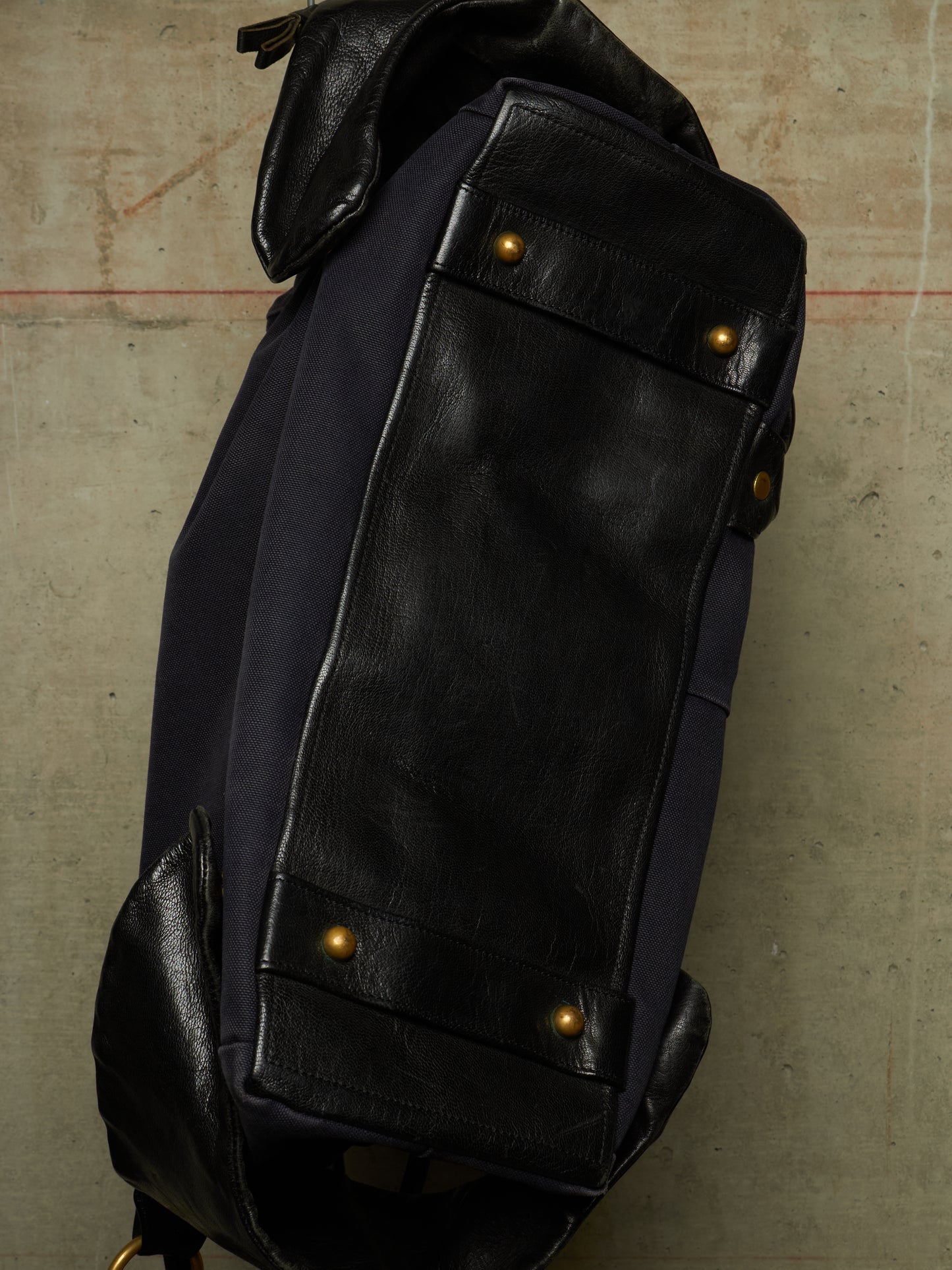 Close-up of a dark navy and black leather backpack corner with brass-tone rivets, photographed against a textured concrete-like surface.