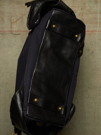 Close-up of a dark navy and black leather backpack corner with brass-tone rivets, photographed against a textured concrete-like surface.