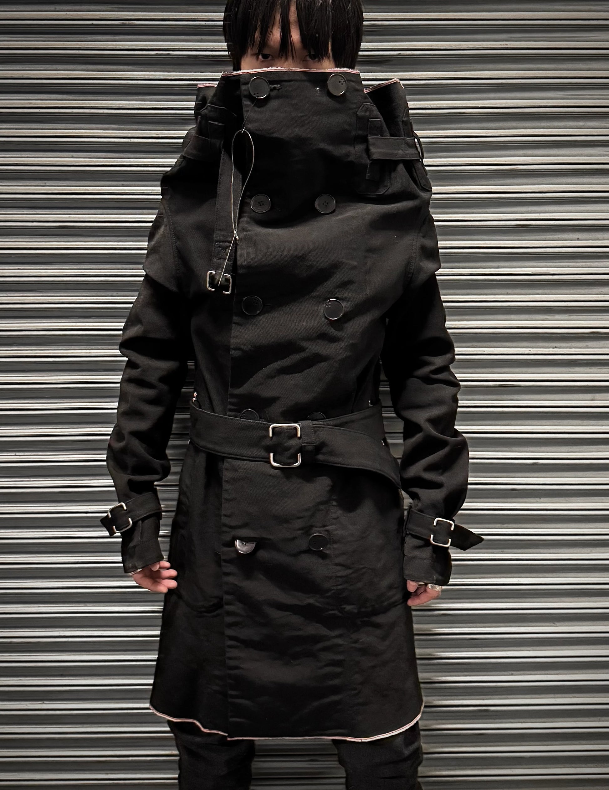 Person wearing a black trench coat with a belt against a metallic background