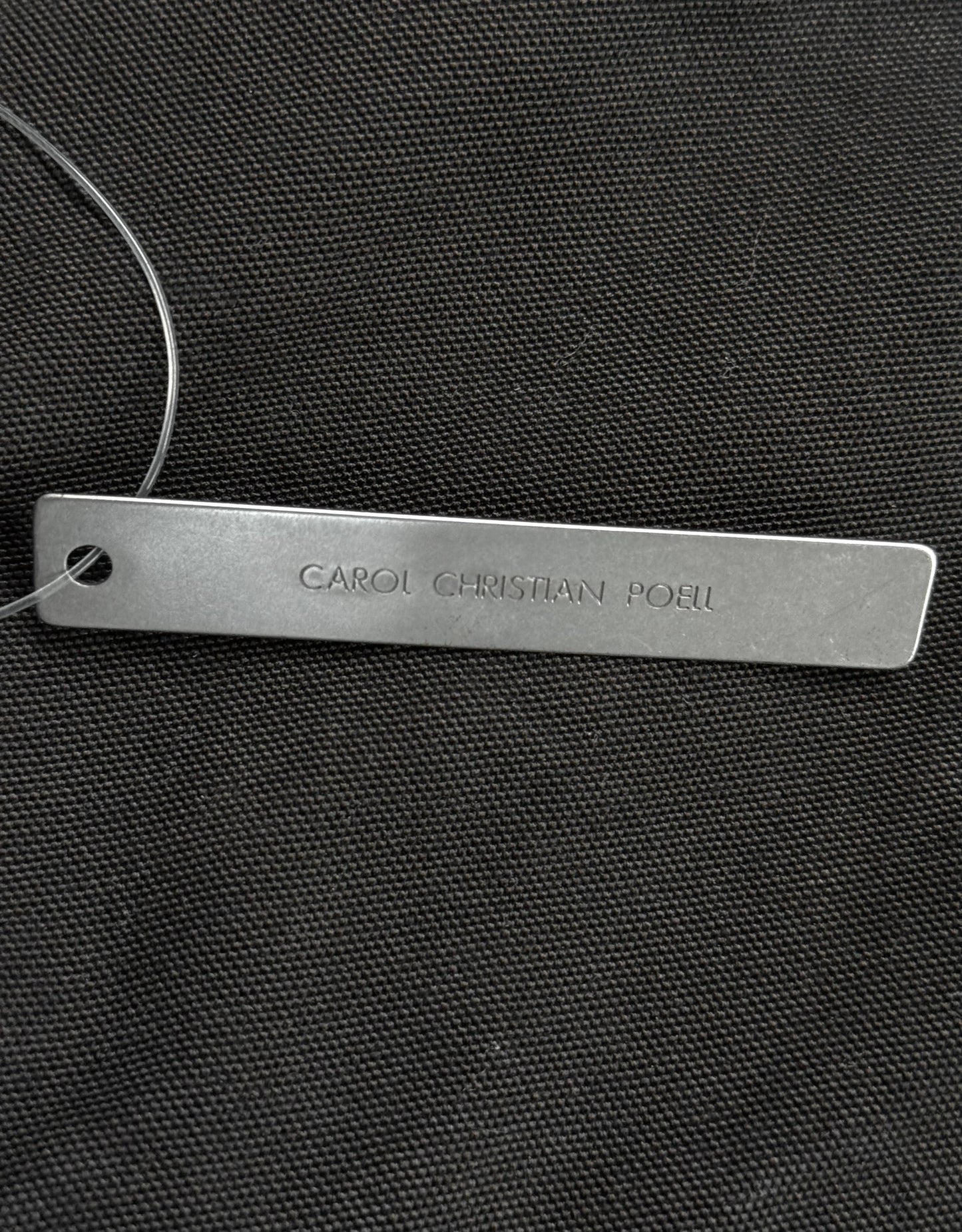 Metal tag with 'CAROL CHRISTIAN POLO' engraved on a black textured background