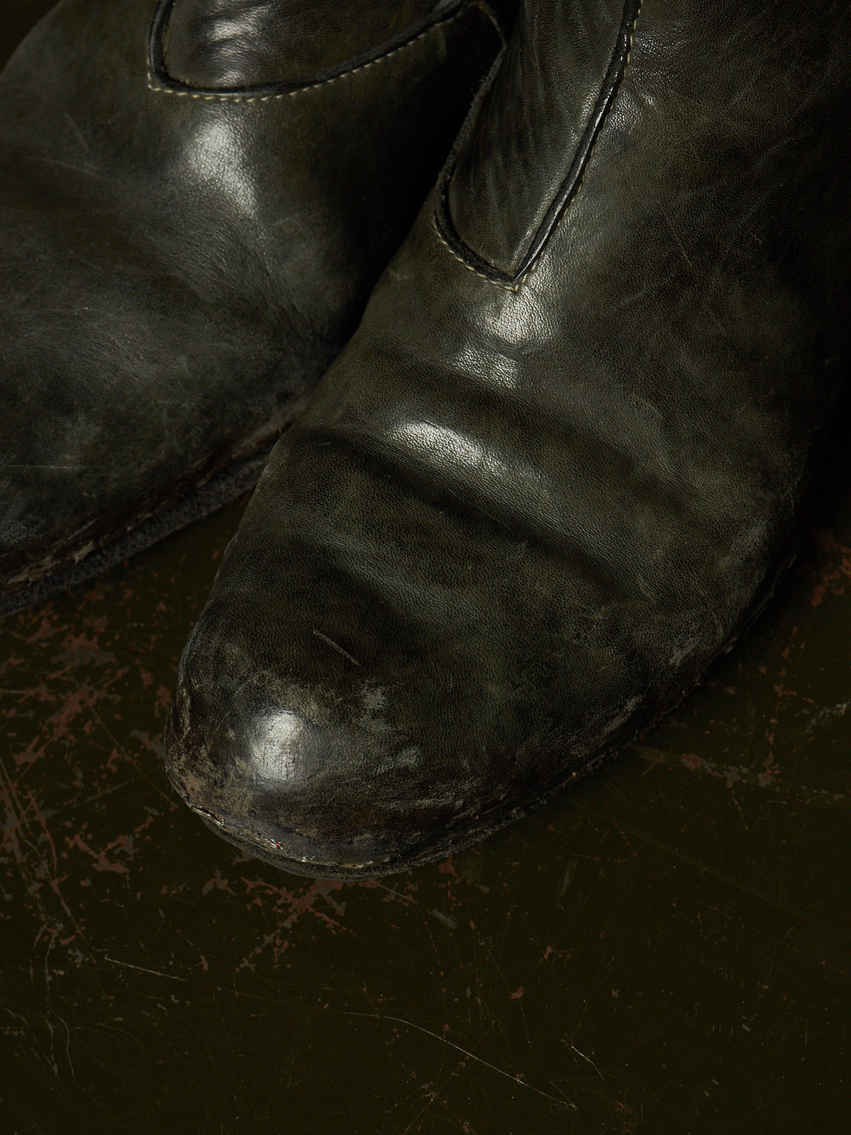 A close-up of a pair of dark distressed leather boots, showing the texture and wear on the toe and vamp, set against a dark, textured background.