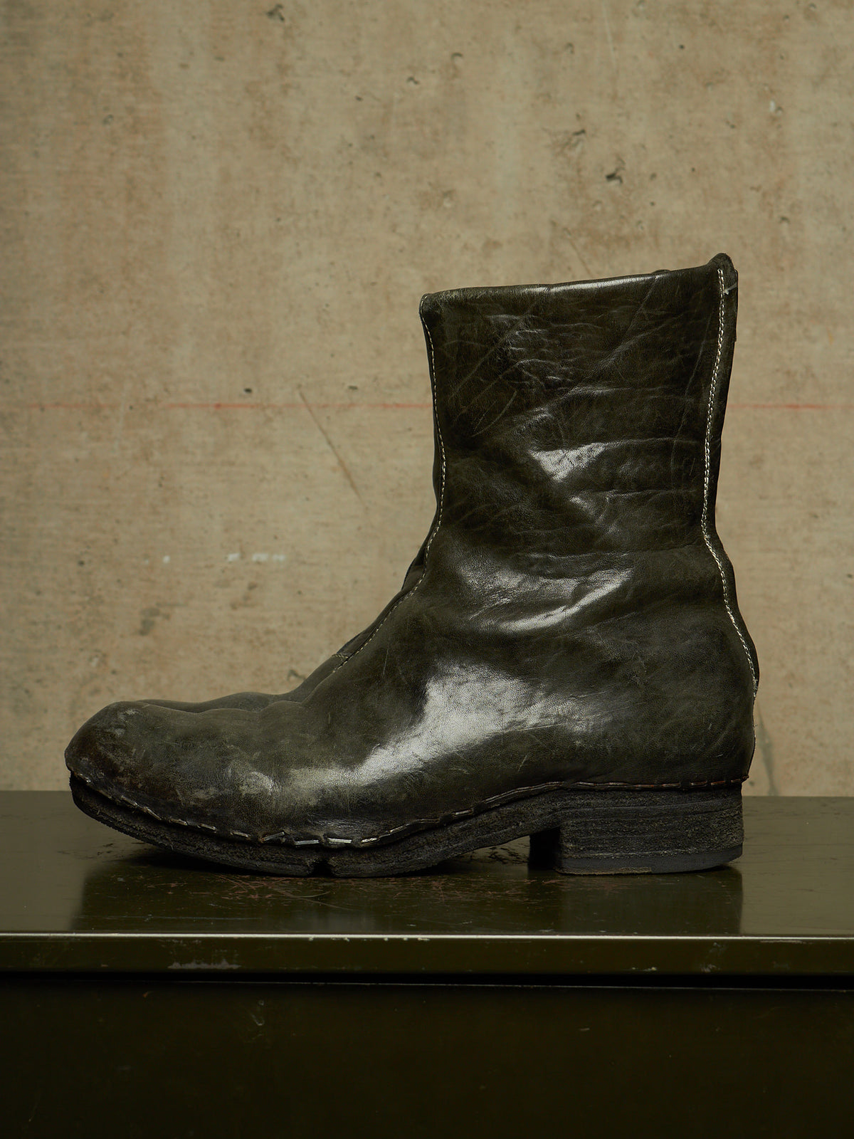A single dark olive green leather ankle boot with a low block heel, displayed in profile on a dark surface against a textured beige wall.