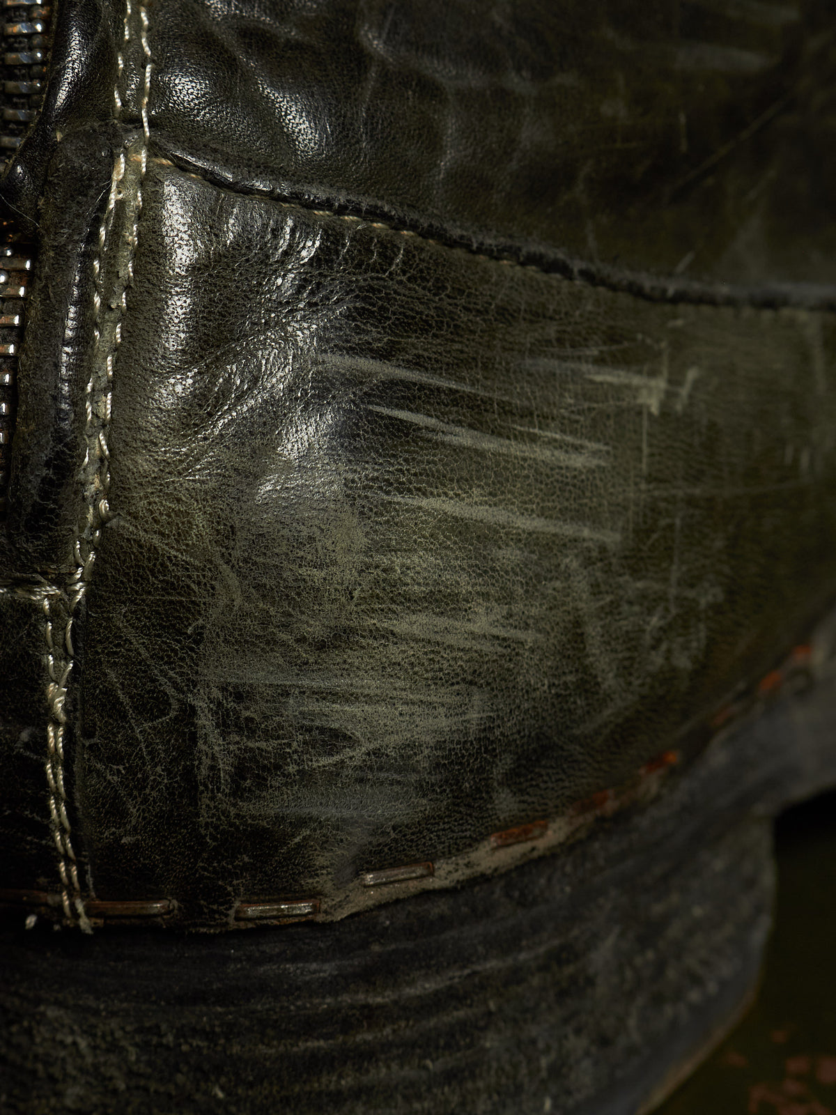 Close-up of a worn dark leather boot showing scuffed texture, heavy scratches, and distressed stitching.