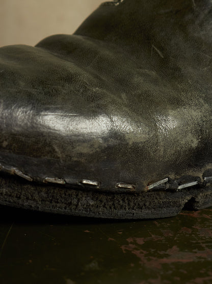 A close-up shot of a dark, heavily distressed leather boot toe showing the textured grain, a worn sole edge with silver metal eyelets, and contrast stitching.